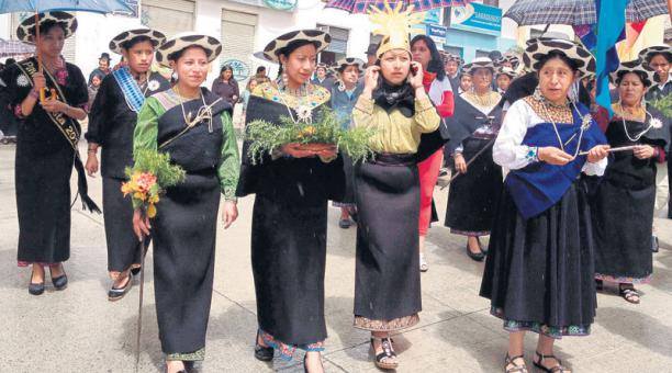 A group of women wearing traditional clothing and wide-brimmed hats participate in a colorful parade, holding plants and objects, showcasing their cultural heritage.