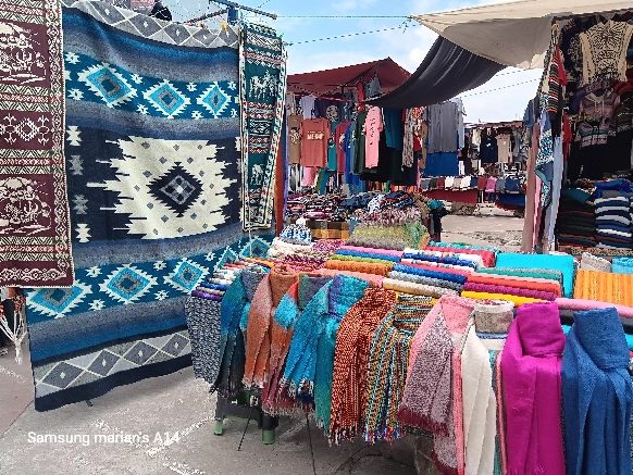 A vibrant marketplace displaying colorful woven textiles, including scarves and patterned blankets, with various clothing items hanging in the background.