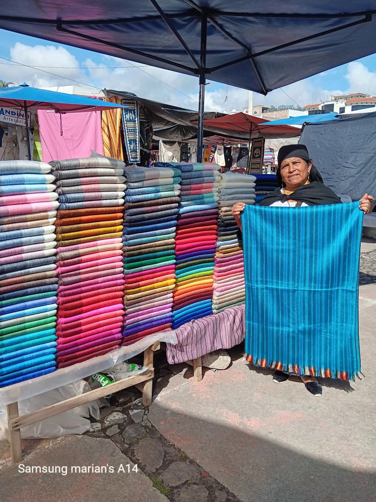 A vendor displays a variety of colorful woven textiles in a market, while holding up a blue striped shawl.