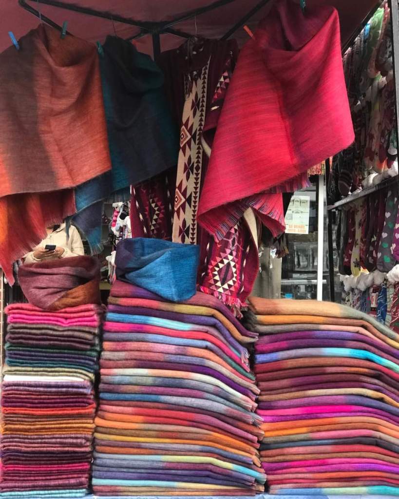 A colorful display of woven textiles hanging on a rack, with a stack of neatly arranged fabrics in various colors and patterns in the foreground.