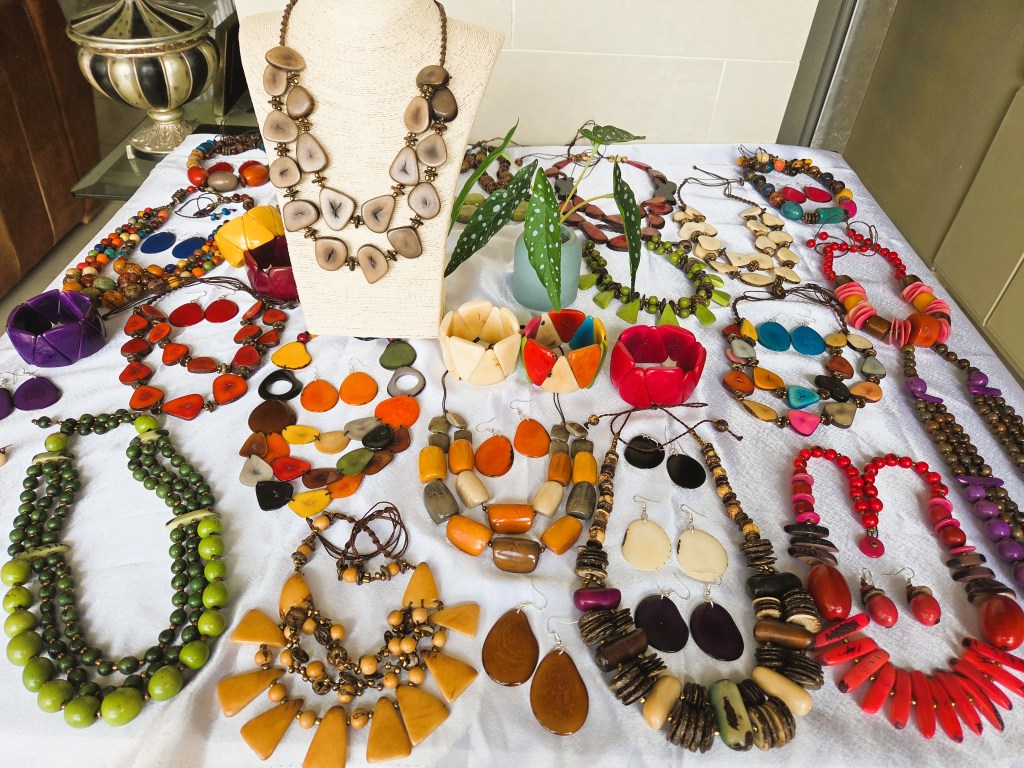 A variety of colorful handmade jewelry displayed on a white tablecloth, including necklaces, earrings, and bracelets made from different materials and designs, with a decorative plant in the background.
